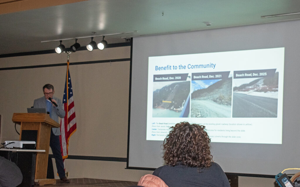 An engineer stands on a stage behind a podium motioning towards a screen behind him that displays a presentation. The presentation shows a side-by-side comparison of a road repair project in Haines resulting from extensive landslide damage with the images going from left to right as the road is fixed.