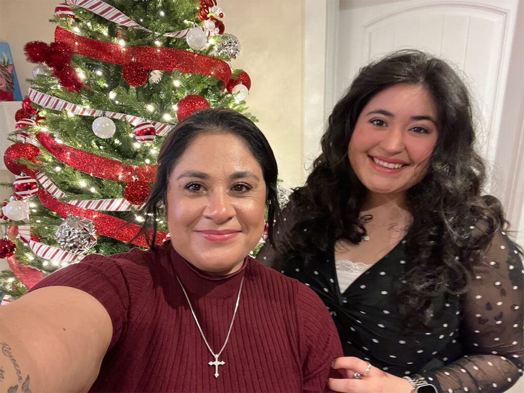 A mother and daughter are standing next to each other smiling. There is a decorated Christmas tree behind them.